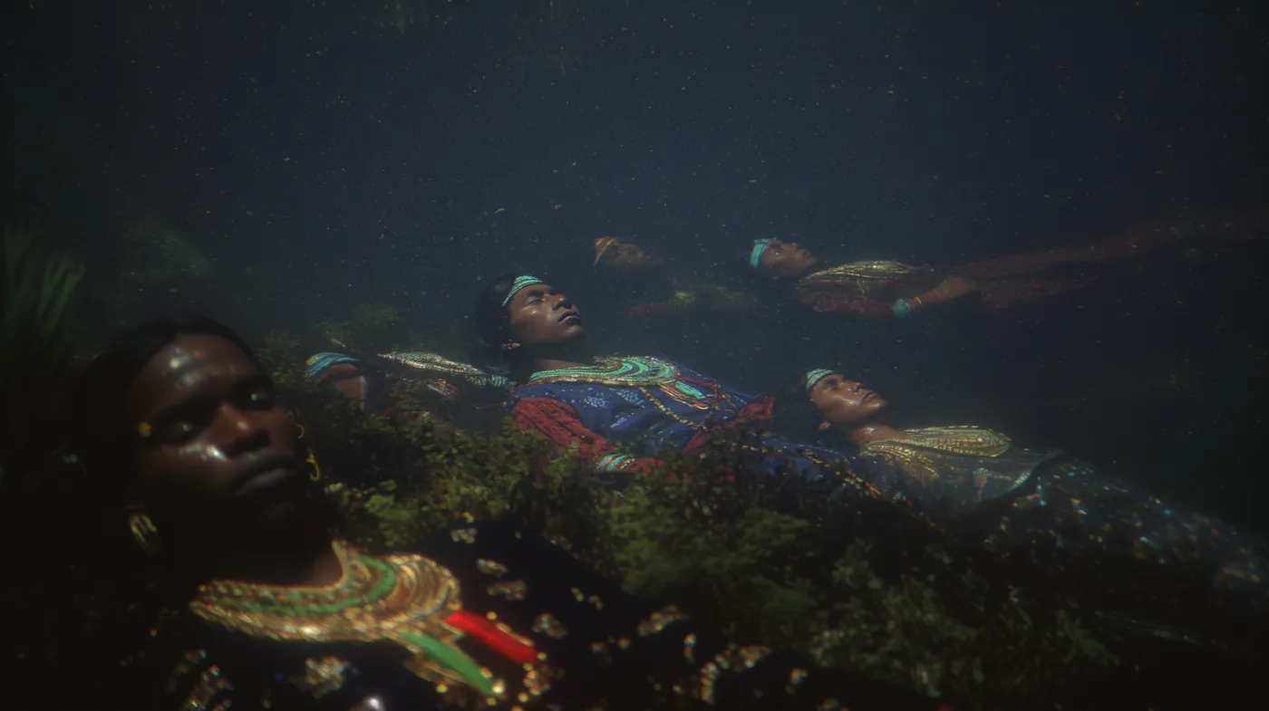 Fotografía submarina en tonos verde oscuro de varias personas flotando bajo el agua con los ojos cerrados; en primer plano, una persona con un collar de cuentas doradas.