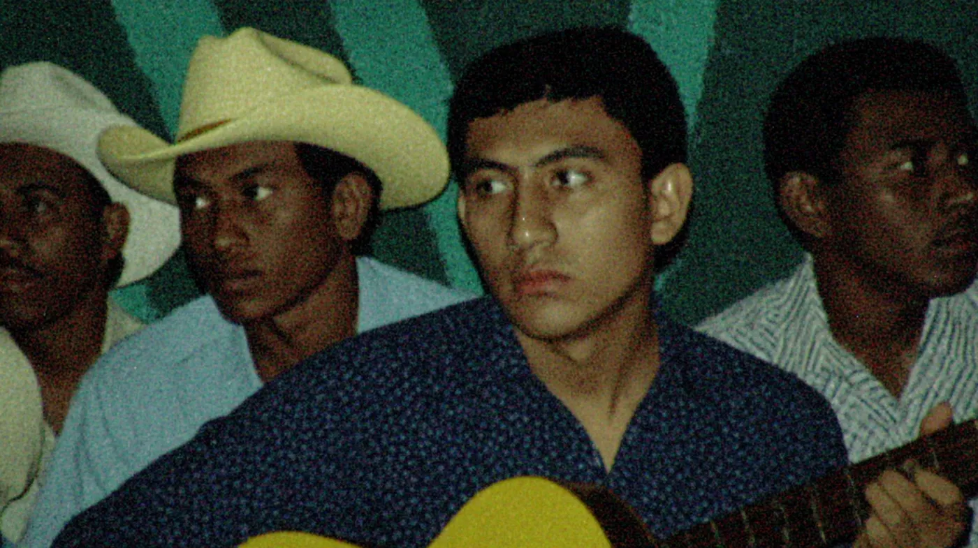Fotografía de estilo vintage de un joven tocando una guitarra acústica en primer plano, con otros hombres con sombreros vaqueros detrás, frente a una pared verde.