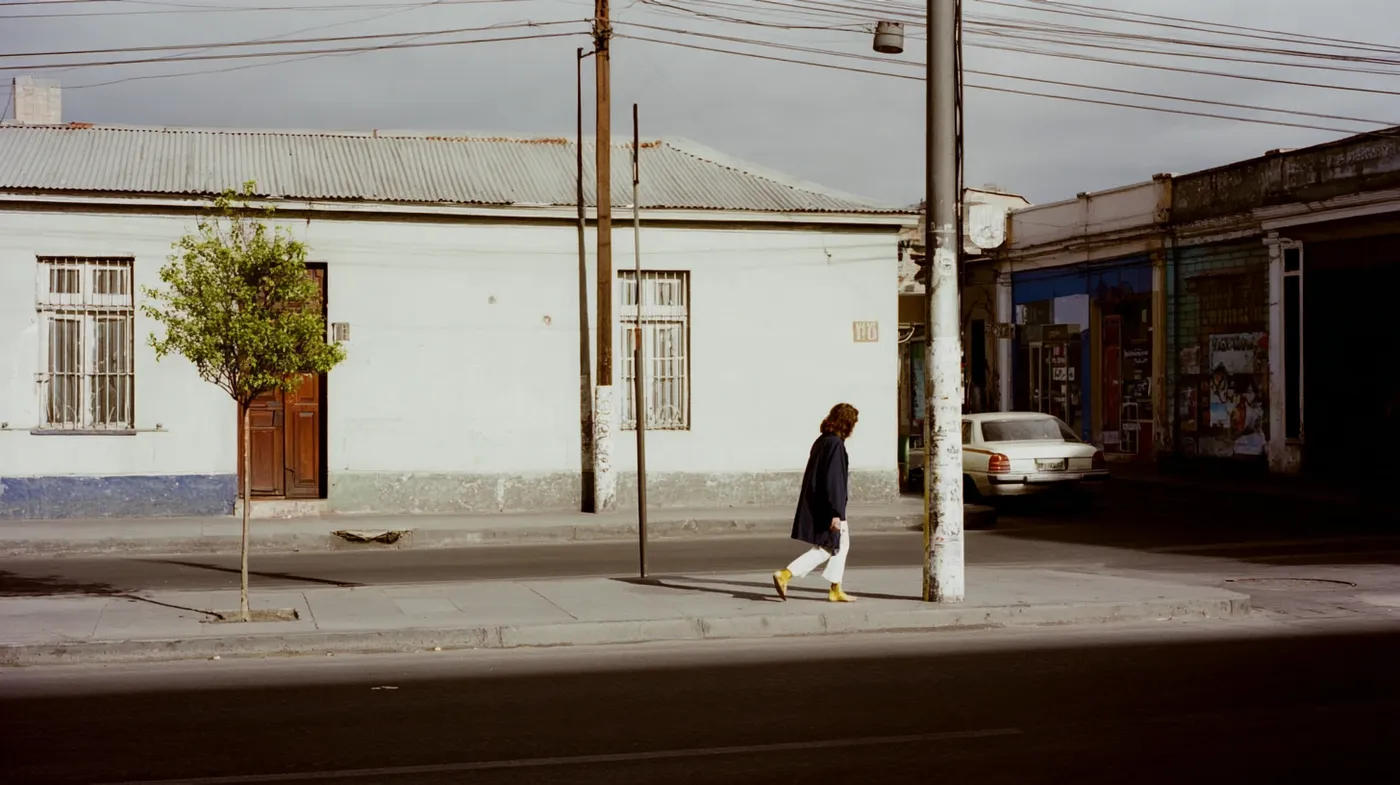 Hombre caminando por la acera de un barrio de casas bajas, junto a un poste con cables eléctricos, un auto estacionado y un local en la esquina, bajo luz de tarde.