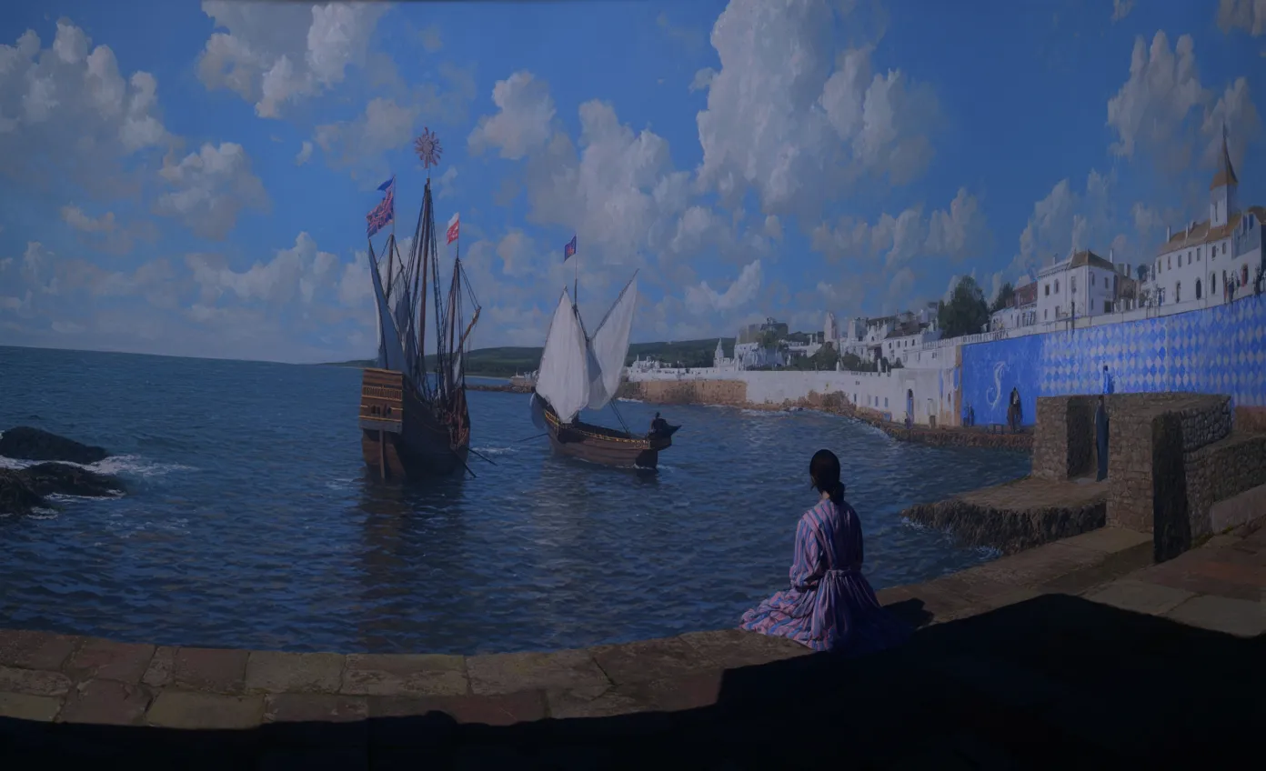 Una mujer con vestido morado está sentada de espaldas sobre un muro de piedra frente a un puerto con dos barcos de vela antiguos y un pueblo de casas blancas, bajo un cielo azul con nubes.